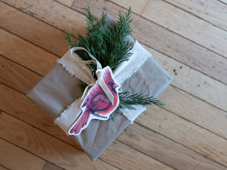 A DIY touch, this gift wrapped in brown paper and burlap ribbon features a cardinal illustration and sprigs of evergreen, evoking the charm of handmade Christmas ornaments. It rests gracefully on a wooden floor.
