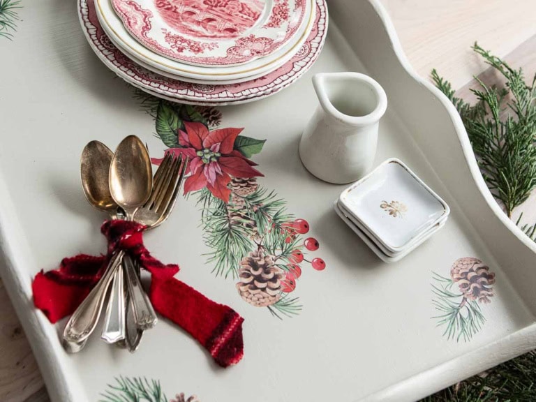 A festive and upcycled serving tray with a holiday-themed design featuring pinecones and poinsettias. It holds red and white plates, a small white pitcher, two decorative dishes, and a bundle of vintage silverware tied with a red ribbon. Pine branches are nearby.