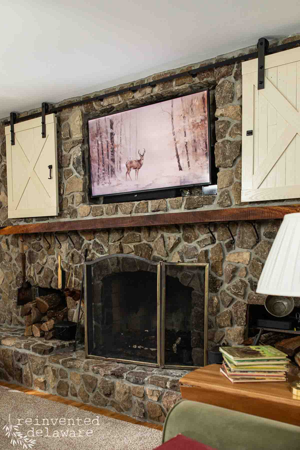 A cozy living room with a stone fireplace adorned with a charming woodland Christmas mantel. A TV above showcases a deer in a snowy forest, framed by barn-style wooden doors. Firewood is stacked to the left, while a small table with books and a lamp sits invitingly in the foreground.