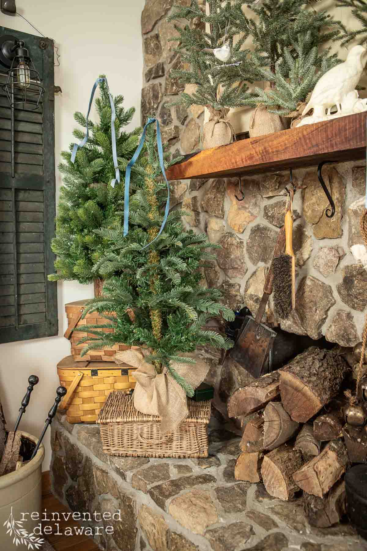 A rustic stone fireplace exudes charm as small evergreen trees in burlap-wrapped baskets grace the woodland Christmas mantel. Stacked firewood and vintage decor enhance the cozy, festive atmosphere, while blue ribbons adorn the trees, completing this enchanting scene.