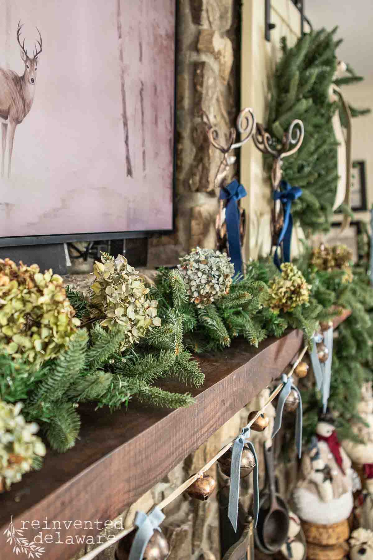 A cozy fireplace mantle decorated for Christmas with pine garland, dried hydrangeas, and ribbons. Above, a painting featuring a deer in a snowy landscape. Two reindeer figurines and a large wreath are visible on the stone wall.