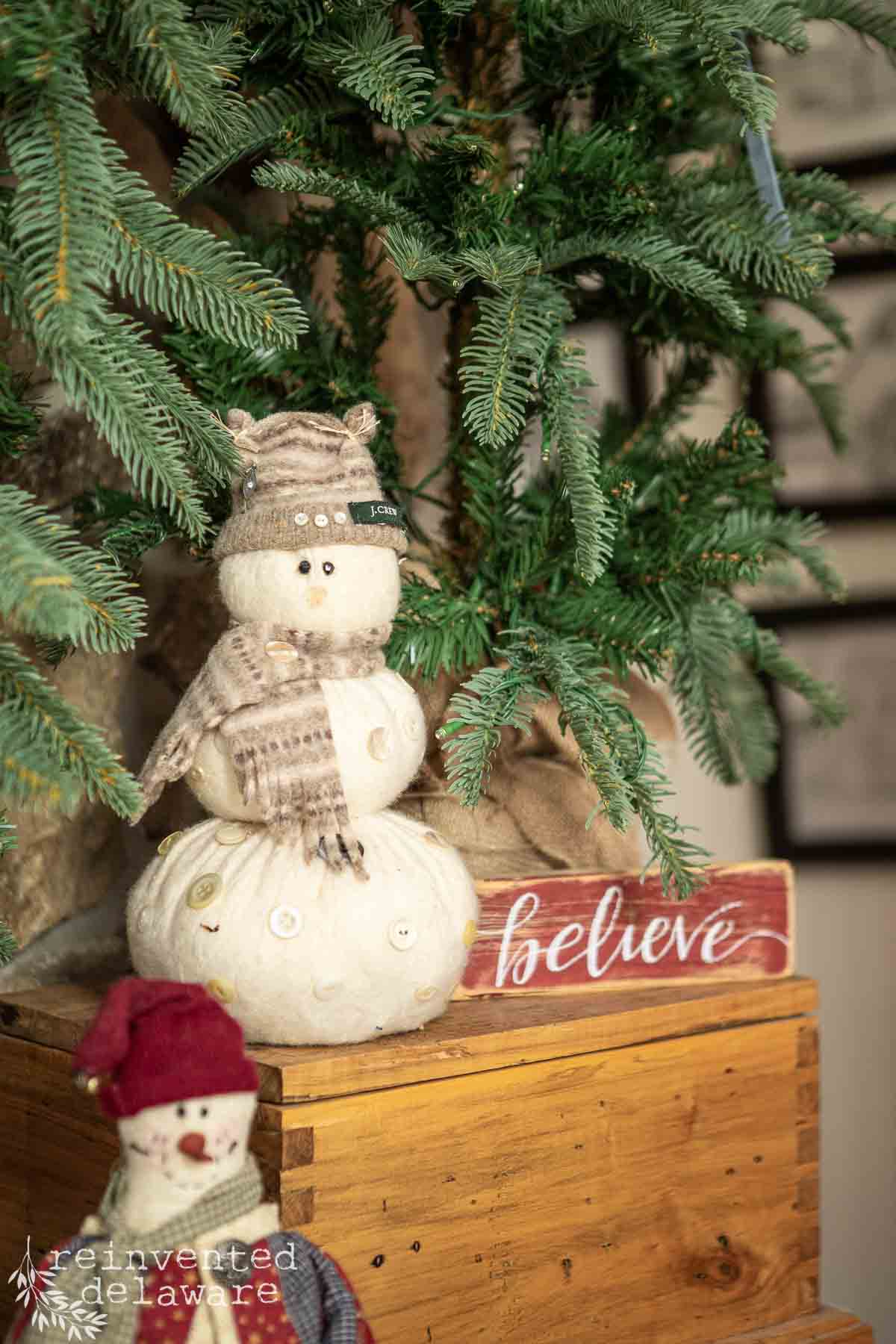 A small snowman figurine in a hat and scarf stands on a woodland Christmas mantel, nestled atop a wooden box beside a "believe" sign. In the background, a larger decorated snowman and the glow of a partially visible Christmas tree add to the festive charm.
