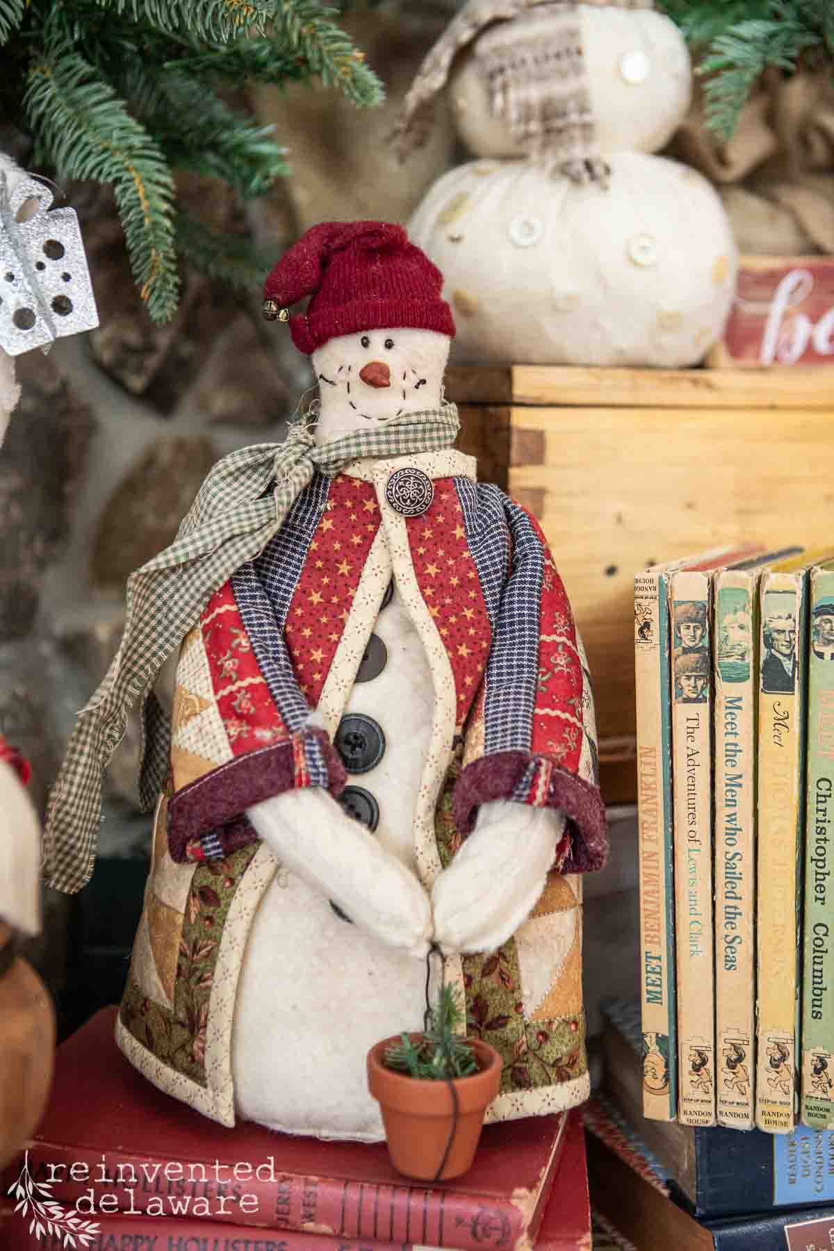 A handmade snowman figurine with a red hat and patchwork coat holds a small potted plant. It stands on the woodland Christmas mantel beside vintage books, surrounded by holiday decor, including a small fabric pumpkin.