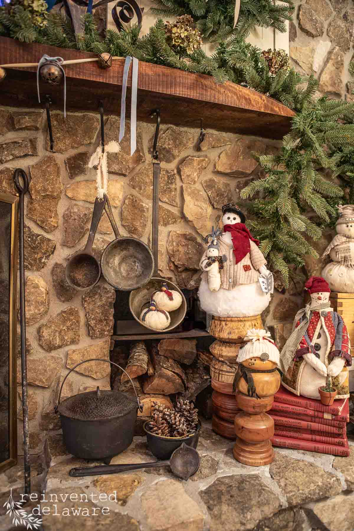 A rustic fireplace hearth decorated for the holidays with snowman figurines, evergreen garlands, and antique kitchenware. Stacks of books and logs add a cozy touch to the warm stone setting.