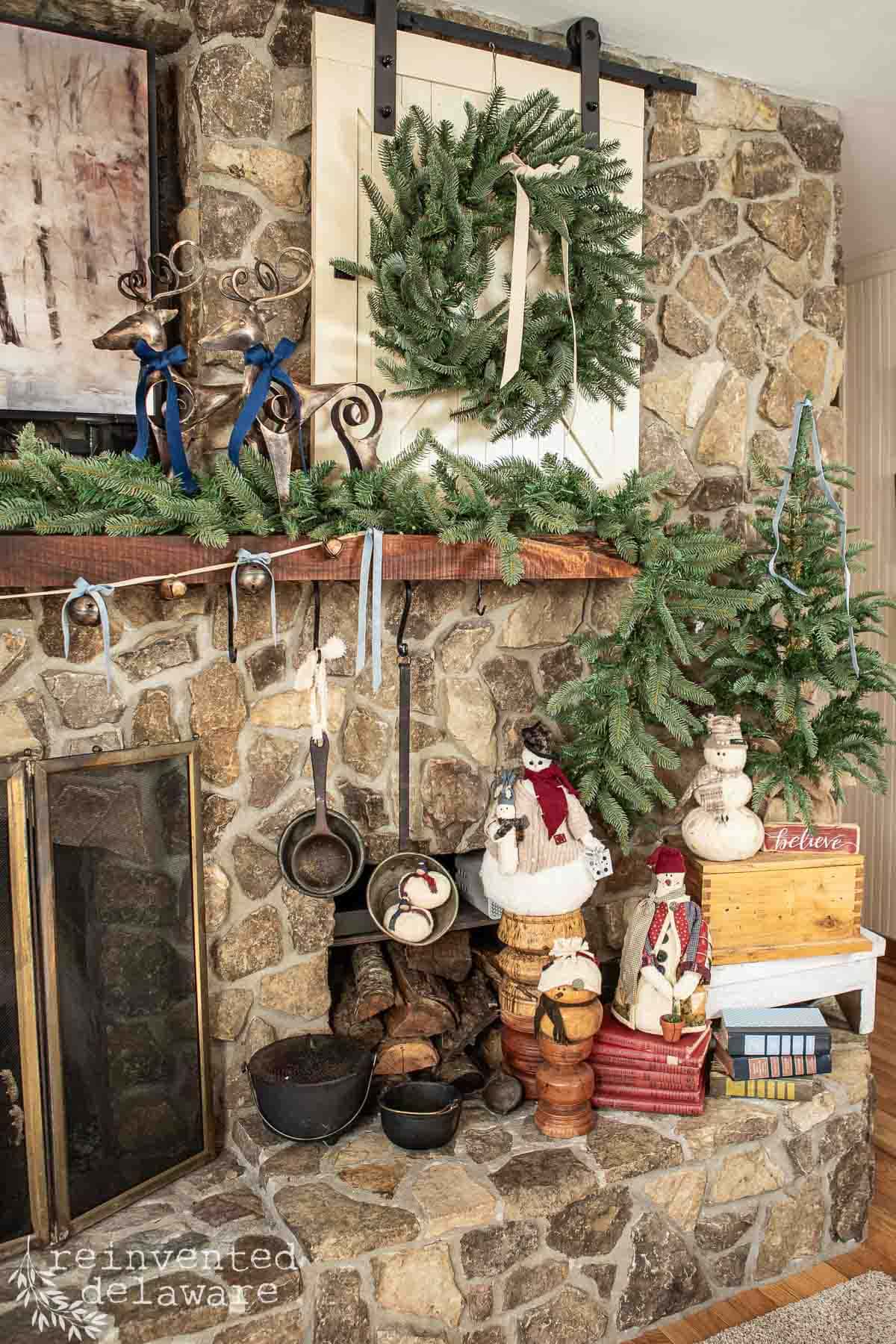A rustic stone fireplace decorated for a woodland Christmas with a wreath, garland, and figurines of snowmen and Santas. The mantel showcases metal candlesticks and wooden accents. Below, a stack of firewood complements the vintage kitchenware's charm.
