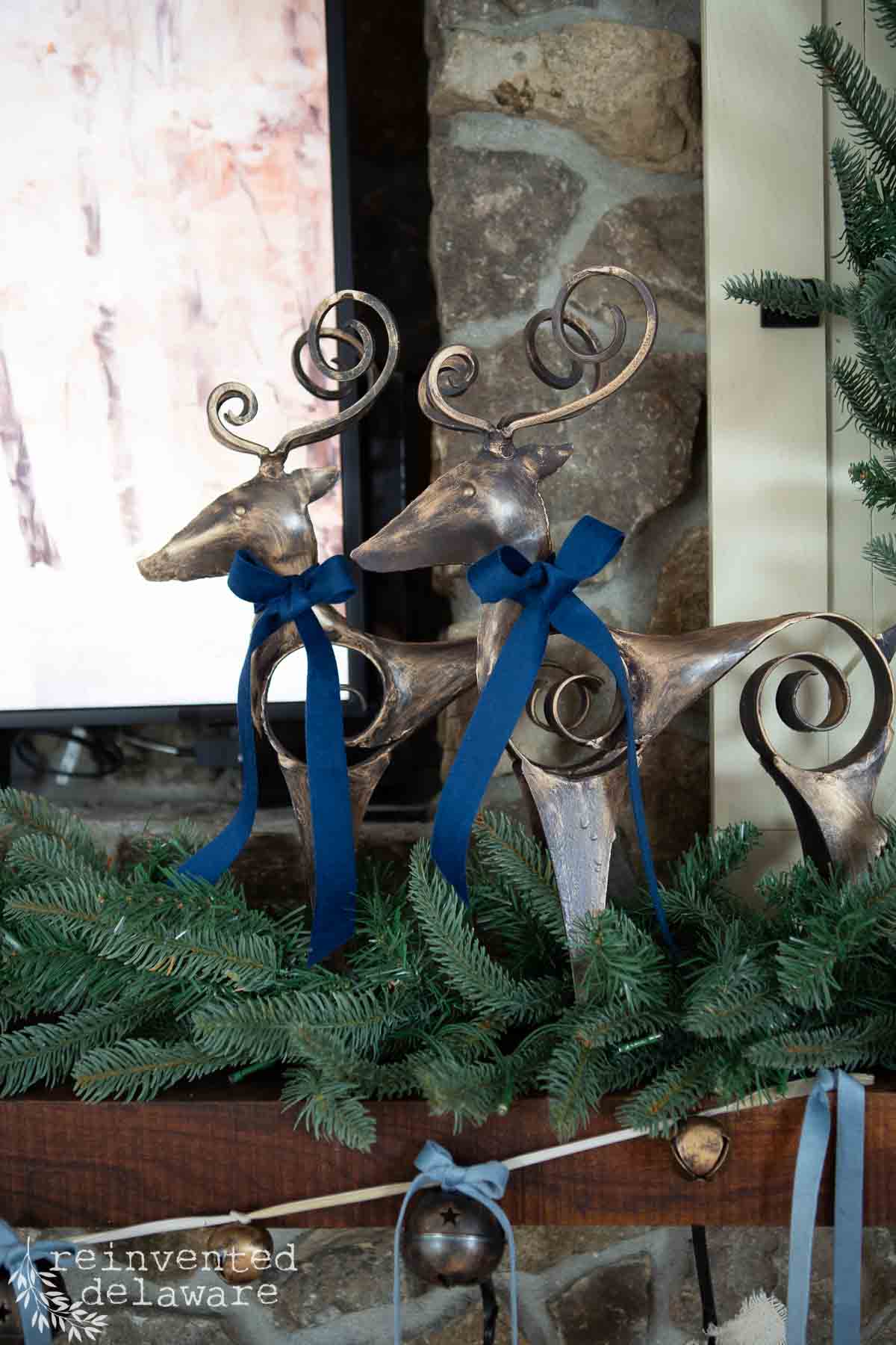 Two decorative metal reindeer with spiral antlers and blue ribbon bows adorn a woodland Christmas mantel, nestled among green fir branches against a stone wall. A wooden surface completes the rustic, festive atmosphere.