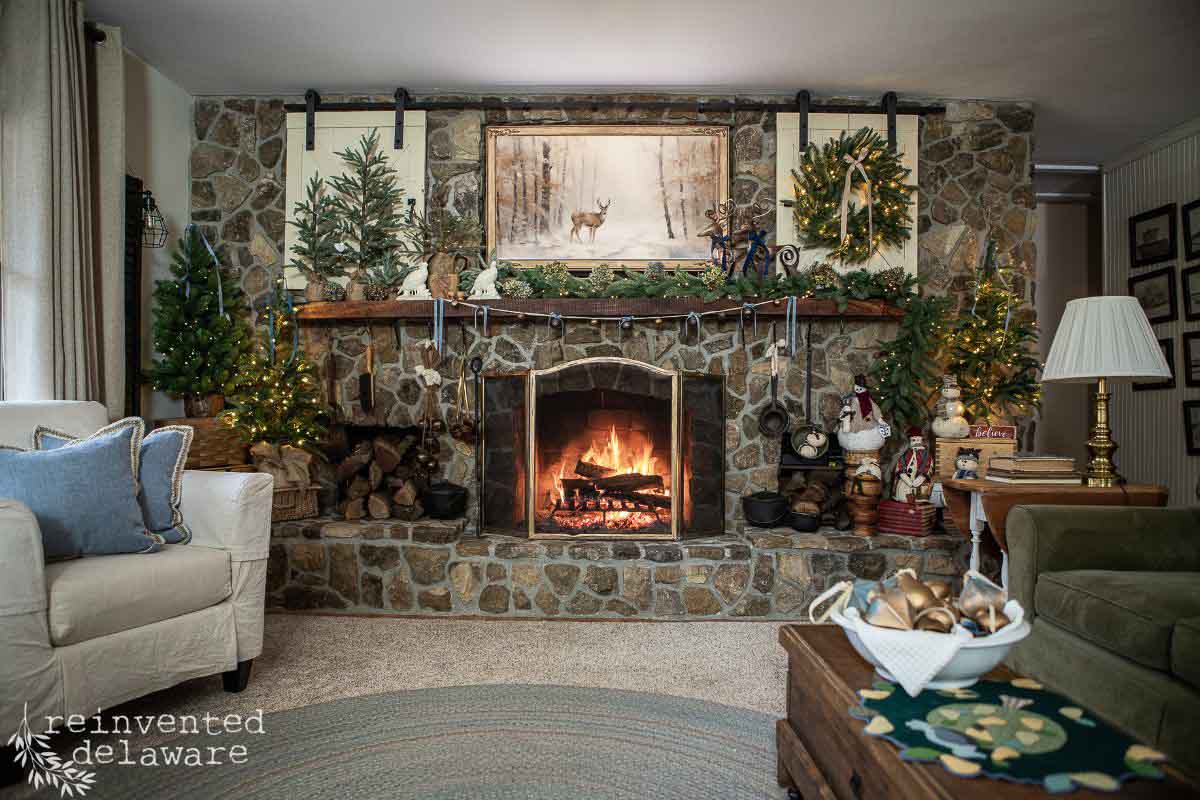 A cozy living room with a stone fireplace, lit fire, and holiday decorations, including wreaths, garland, and mini Christmas trees. A soft chair, side table with lamp, and a coffee table with a tray of decor complete the scene.