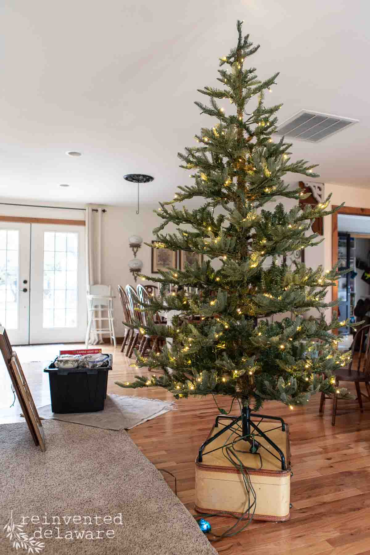 A sparsely decorated Christmas tree with lights stands in a living room with wooden floors. A black storage box sits nearby. The room has white walls, a double glass door, and chairs around a table in the background.