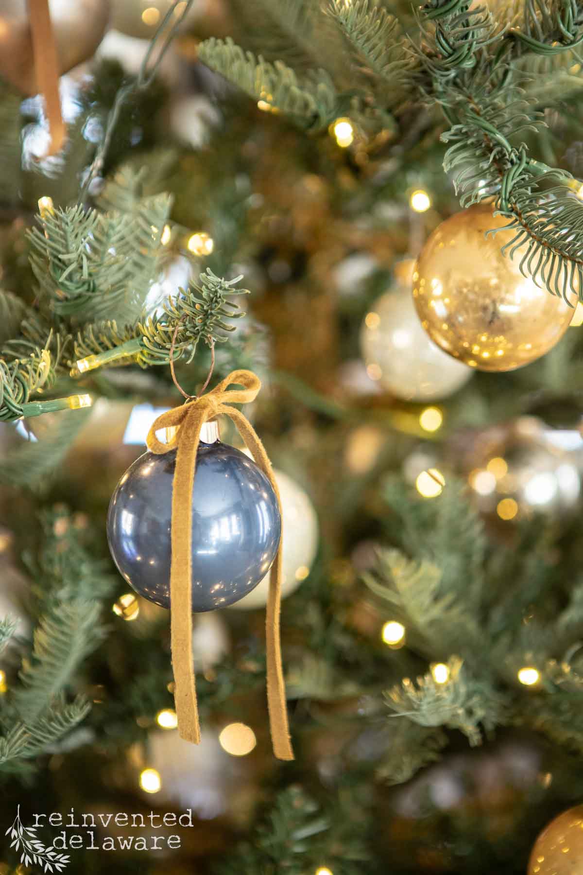 A close-up of a Christmas tree adorned with golden lights and various ornaments, including a prominent blue bauble with a gold bow. The tree branches are lush and green. The words "reinvented delaware" are visible in the bottom left corner.