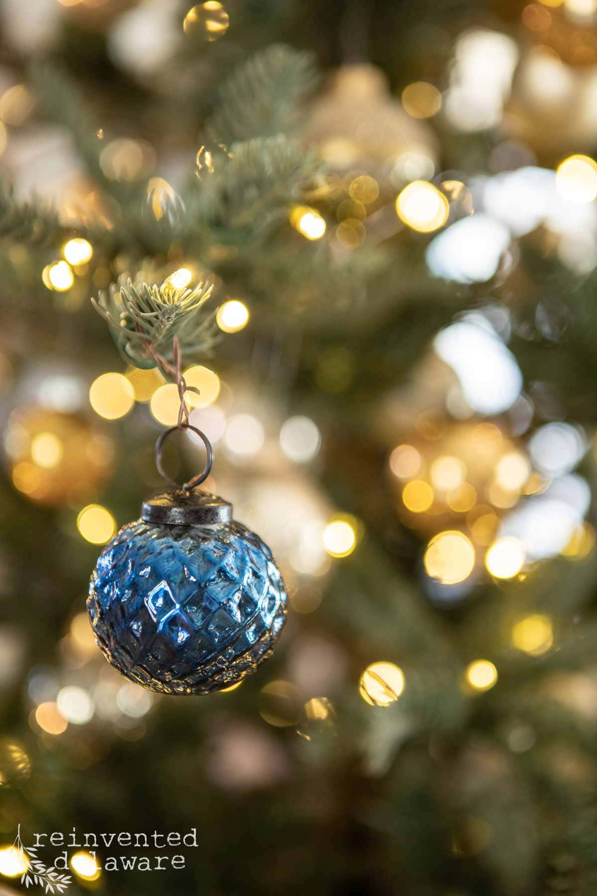 A sparkling blue ornament hangs from a Christmas tree branch, surrounded by blurred, glowing lights.