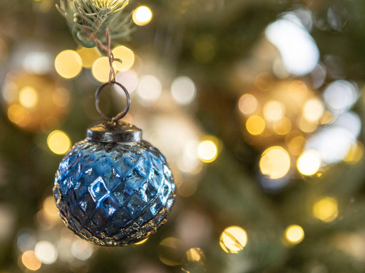 A close-up of a blue glass ornament hanging from a Christmas tree. The background features soft-focus warm yellow and white lights, creating a festive and cozy atmosphere.
