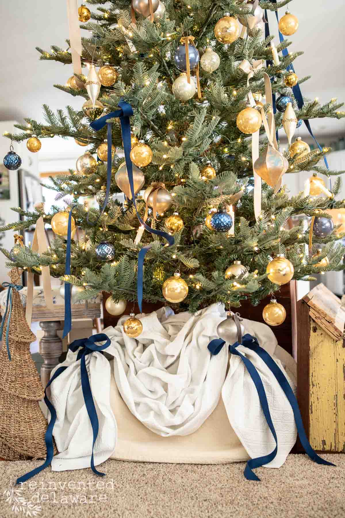A decorated Christmas tree with gold and blue ornaments, blue ribbons, and white fabric at the base. The tree is set in a cozy room with soft lighting, featuring small wrapped presents nearby.