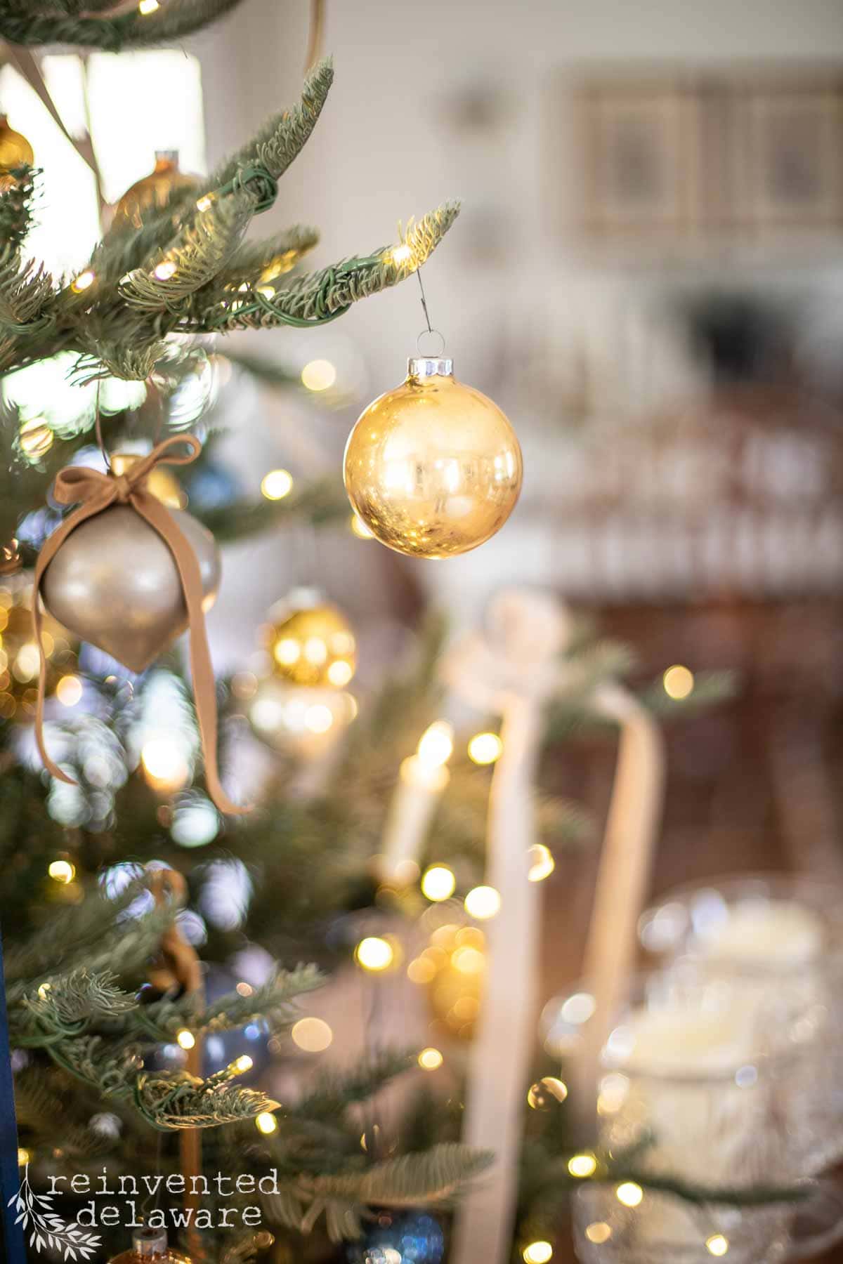 A close-up of a Christmas tree adorned with golden ornaments and warm white lights. The blurred background adds a cozy ambiance, highlighting the festive decorations. The image includes a watermark: "reinvented delaware.