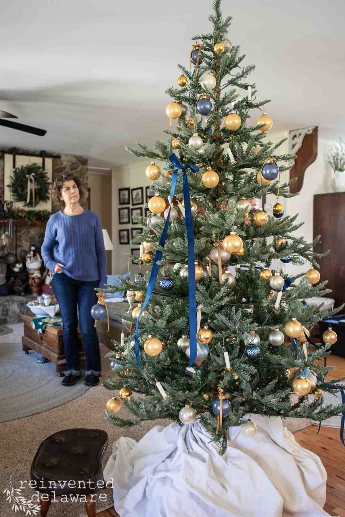 A woman in a blue sweater stands next to a decorated Christmas tree adorned with gold and blue ornaments and ribbons. The tree is set up in a living room with a cozy interior visible in the background.