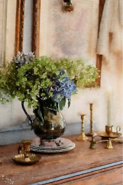 A decorative table setting with a vintage vase holding green and blue hydrangeas. There are brass candlesticks and small brass dishes scattered on the wooden surface. Framed artworks hang in the blurred background.