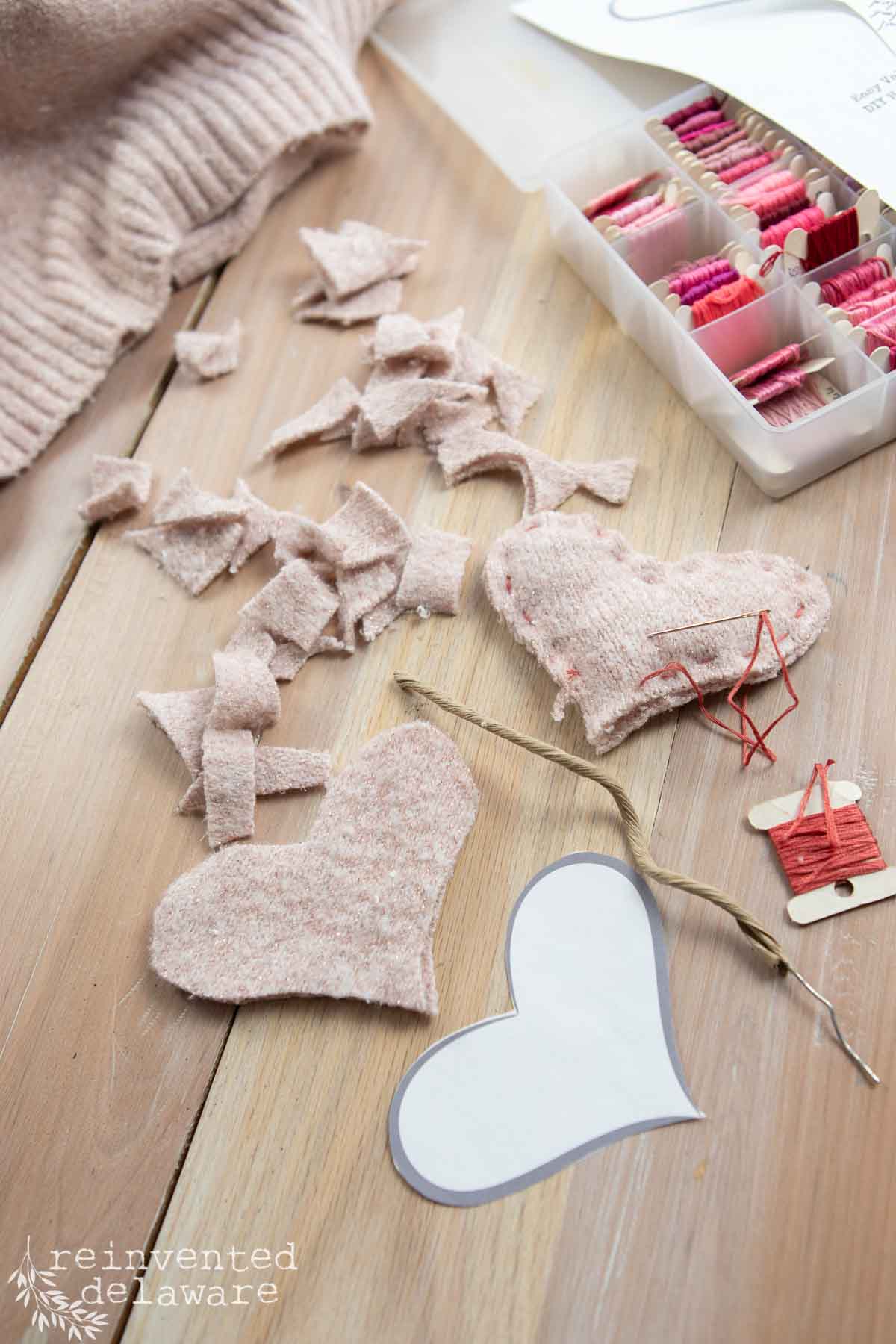 Crafting materials arranged on a wooden table, including stitched fabric hearts, small fabric strips, embroidery floss in various pink shades, and a heart-shaped template. A spool with a needle and red thread is also visible.