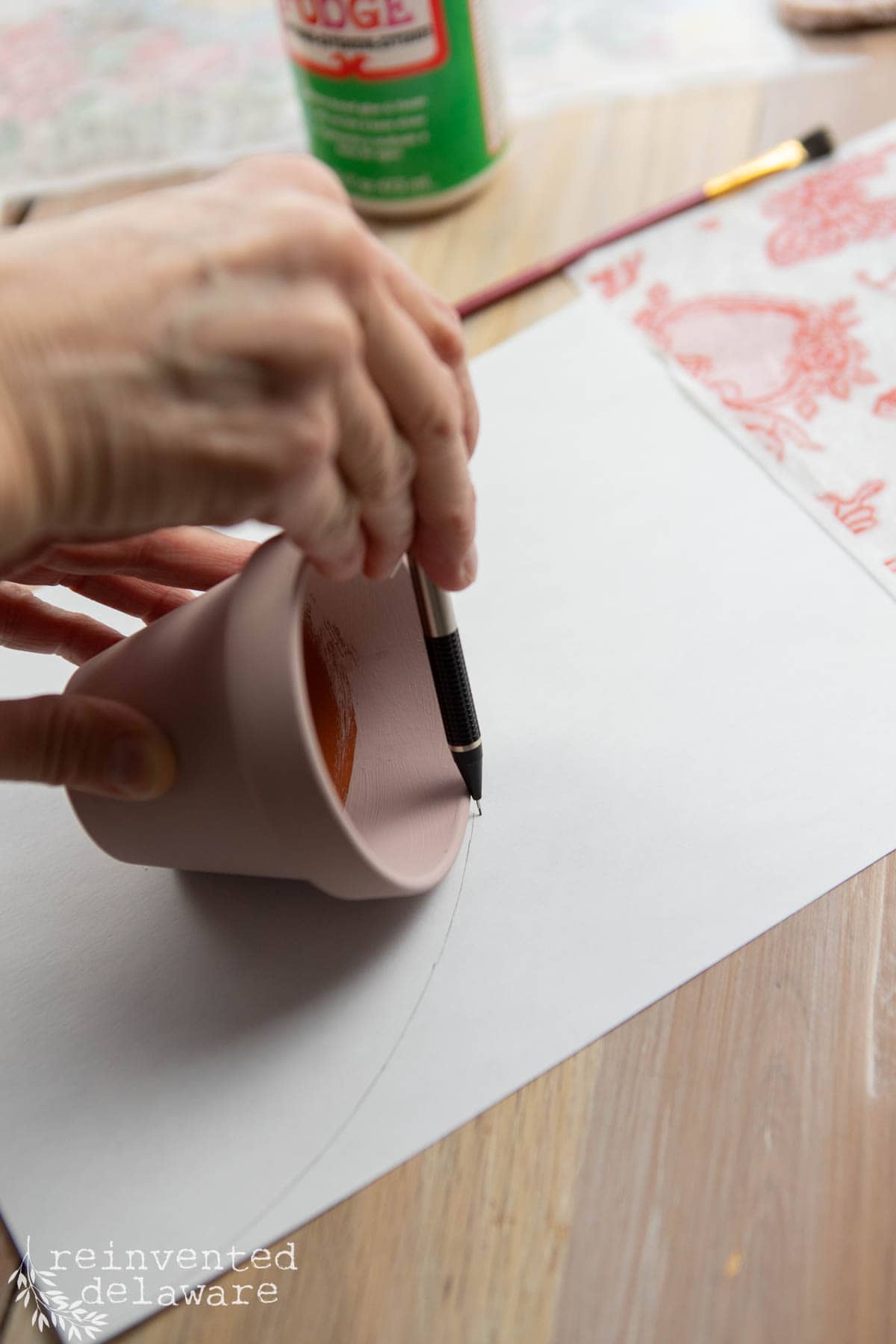 A person uses a pen to trace the outline of a small terracotta pot on white paper. Nearby, a paintbrush and a bottle of Mod Podge hint at an upcoming Valentine's Day decoration DIY project. The focus is on the hands and the pot.
