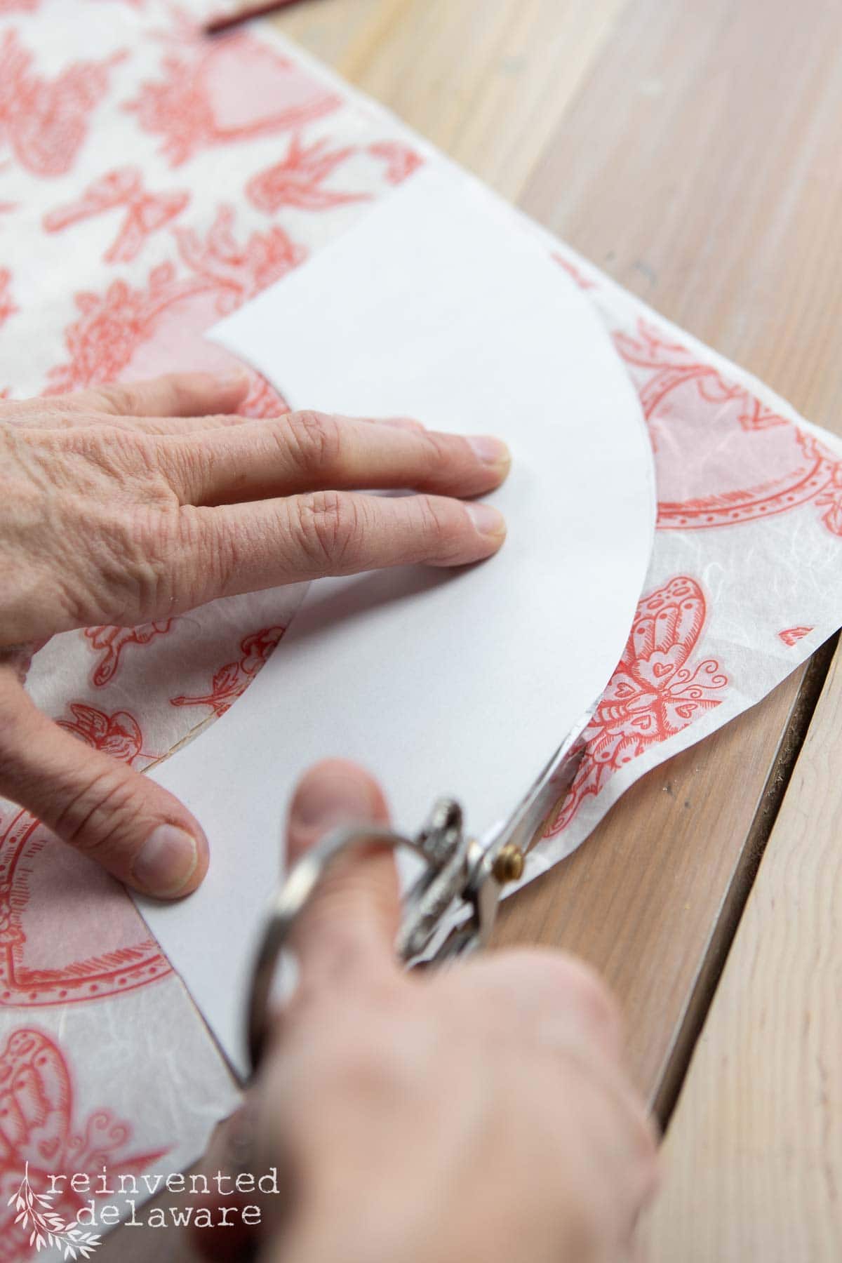 Close-up of hands using scissors to cut along a curved line on a piece of white paper, perfect for crafting Valentine's Day DIY decorations, over red patterned fabric. The fabric rests on a wooden surface, adding a charming touch to your heartfelt creations.