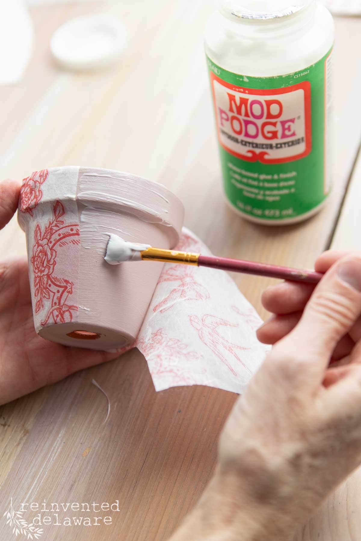 A person applies Mod Podge with a brush to a small pink flower pot, creating the perfect Valentine's Day DIY decor. Decoupage paper with red floral patterns is partially adhered to the pot, while a jar of Mod Podge sits nearby on the wooden surface.