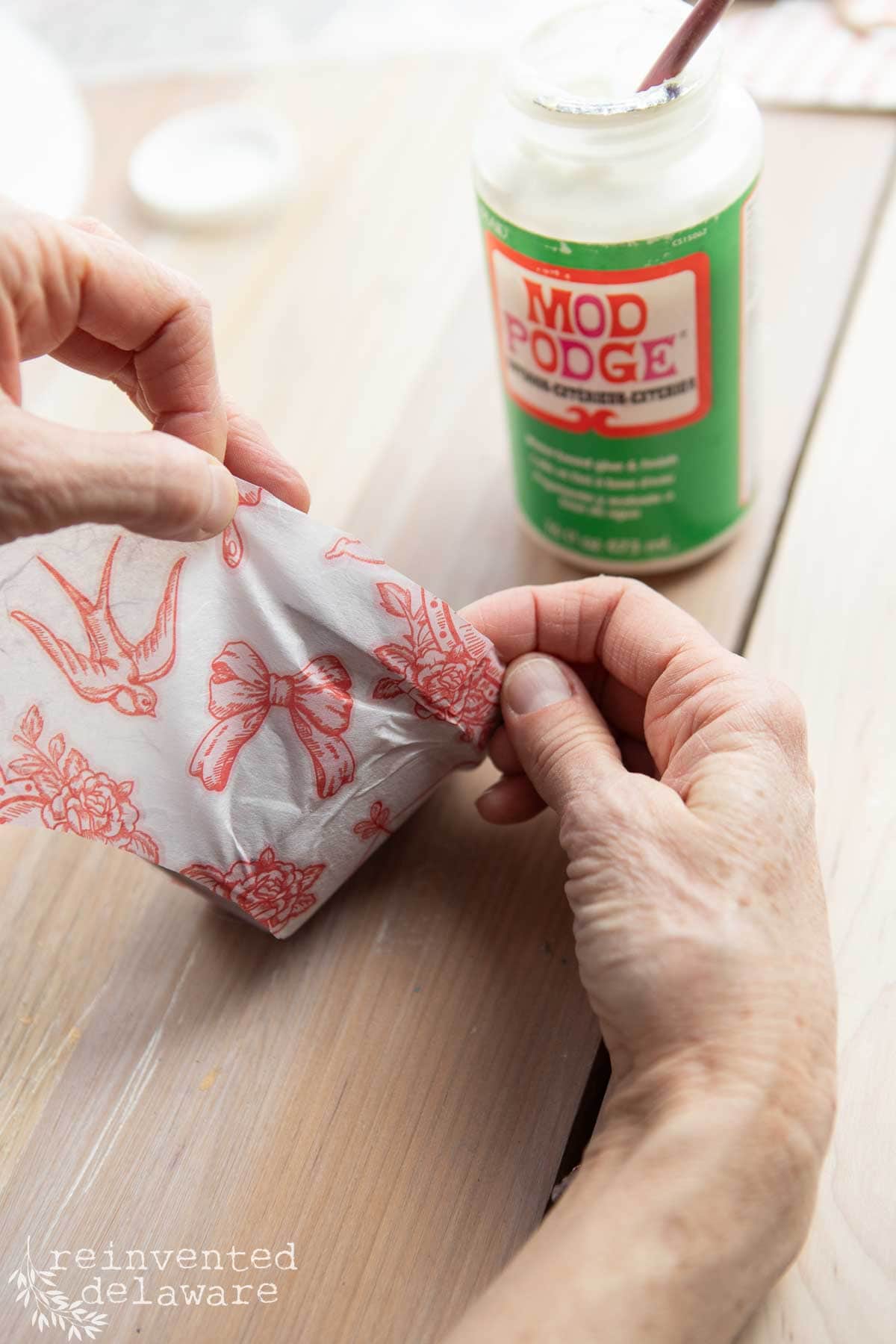 Hands applying Mod Podge to a white surface adorned with red designs of birds, butterflies, and flowers—perfect for a Valentine's decor DIY project. A jar of Mod Podge and a brush sit in the background on a wooden surface, ready to transform decoupage paper into art.