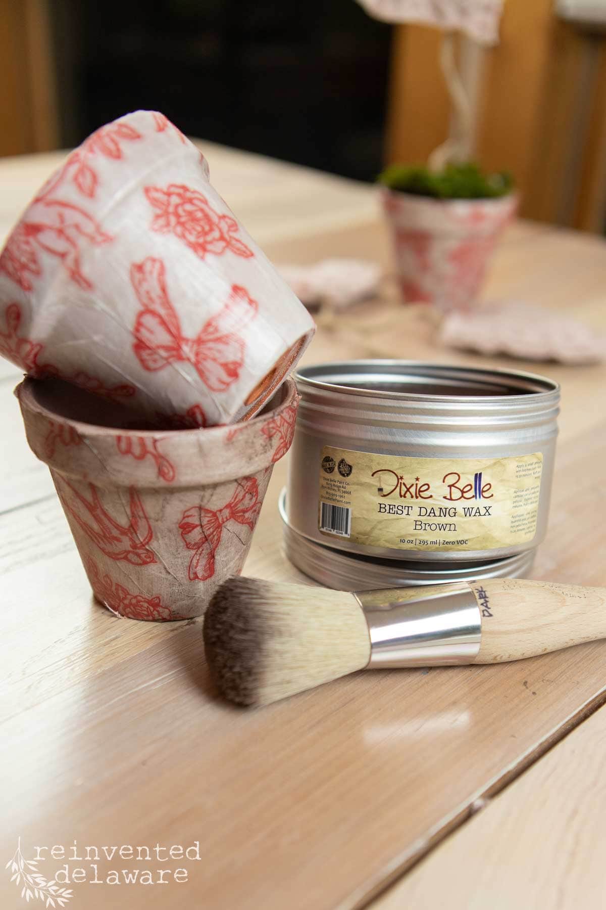 Craft supplies on a table include a paintbrush, a can of Dixie Belle Best Dang Wax in brown, and two terra cotta pots with red butterfly designs. The background is softly blurred, hinting at Valentine’s Day-inspired creativity.