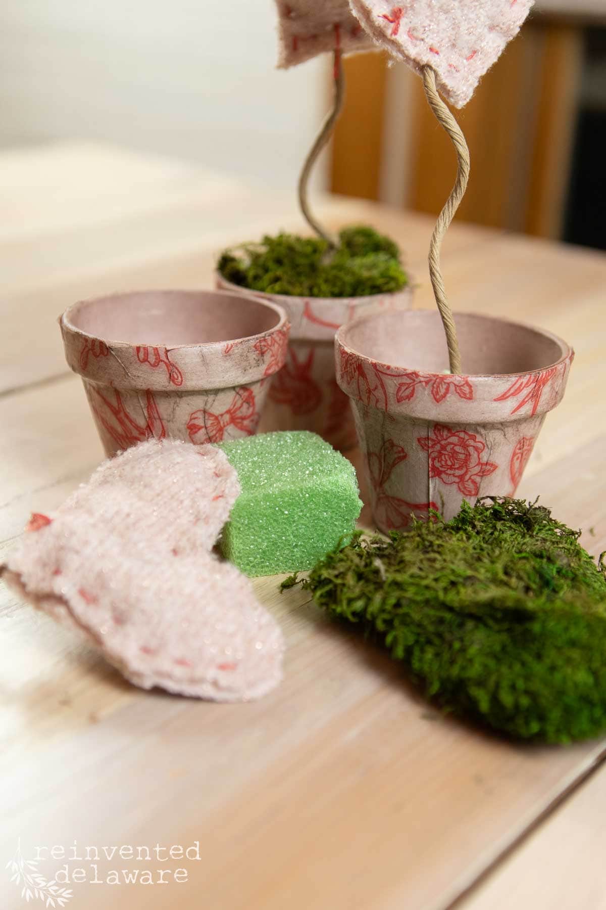 Three small pots covered in pink decoupage paper with red floral patterns are on a wooden table. Two green sponges and pieces of moss are nearby. Two stuffed hearts are attached to flexible wires in the pots for a valentines day diy decor.