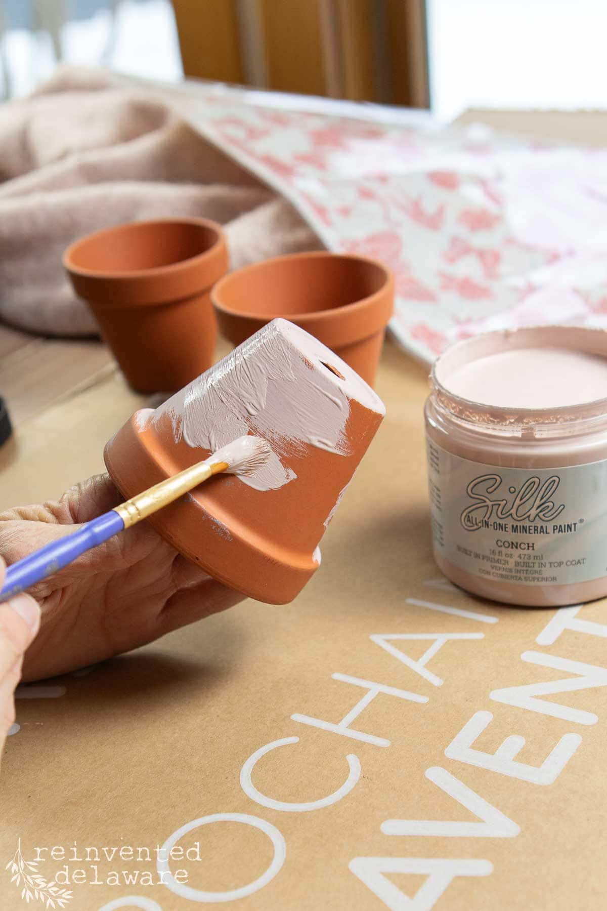 A hand painting a small terracotta pot with pink mineral paint using a brush. The paint jar is labeled "Silk All-In-One Mineral Paint - Conch." Perfect for a DIY Valentine's decor project, two unpainted pots await transformation on the craft table in the background.