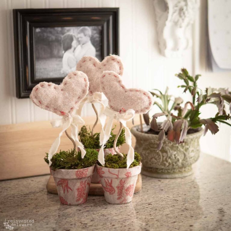 Three small terracotta pots decorated with red patterns hold heart-shaped fabric decorations on sticks with ribbons. The pots are placed on a granite countertop near a green potted plant and a blurred black-and-white photo on the wall.