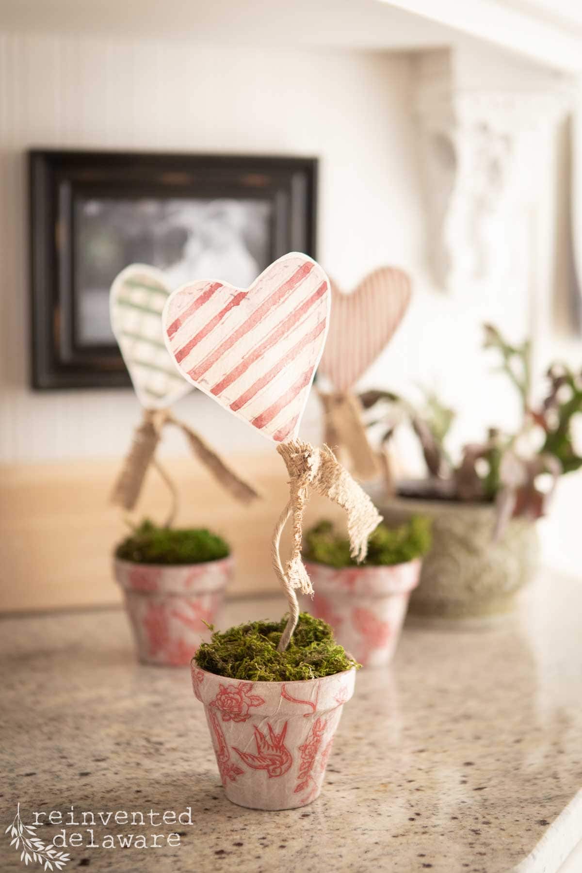 Three small decorative pots sit on a countertop, each with a paper heart on a stem. The hearts, perfect for cute DIY Valentine's Day decor, feature red and white stripes and burlap bows. Moss covers the soil, and a logo for "Reinvented Delaware" is in the bottom left corner.