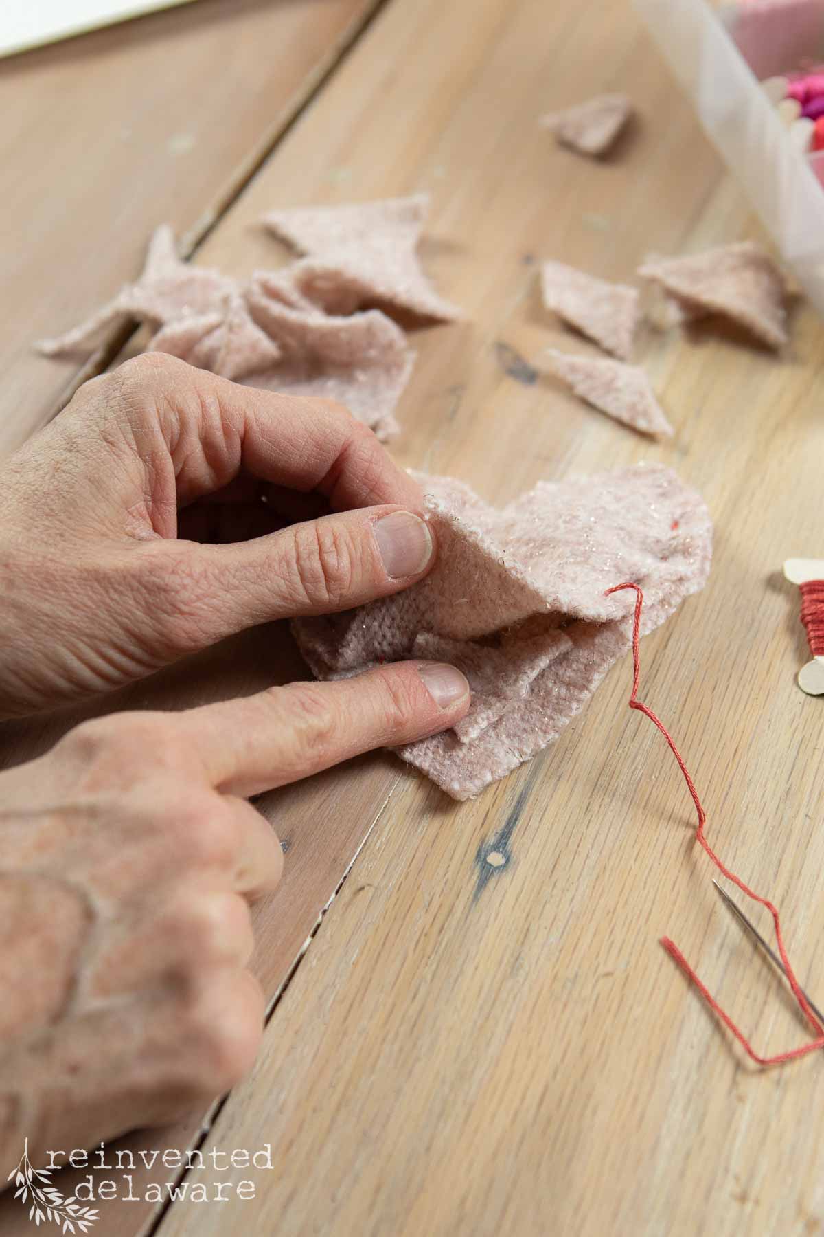 Crafting DIY table decor for Valentine's Day, a pair of hands meticulously sew pink fabric pieces with red thread on a wooden table.