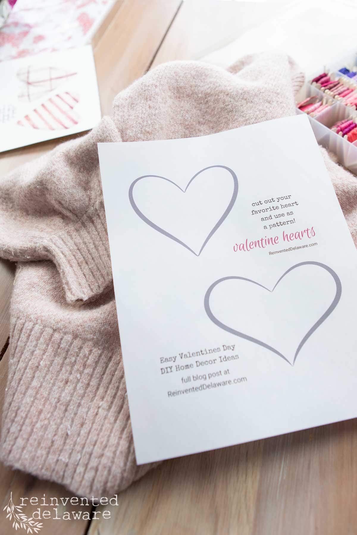 A sheet of paper with outlines of two hearts labeled "valentine hearts" is placed on a knitted pink sweater. Colored embroidery floss is nearby. Text reads "Easy Valentine's Day DIY Home Decor Ideas" and references ReinventedDelaware.com.