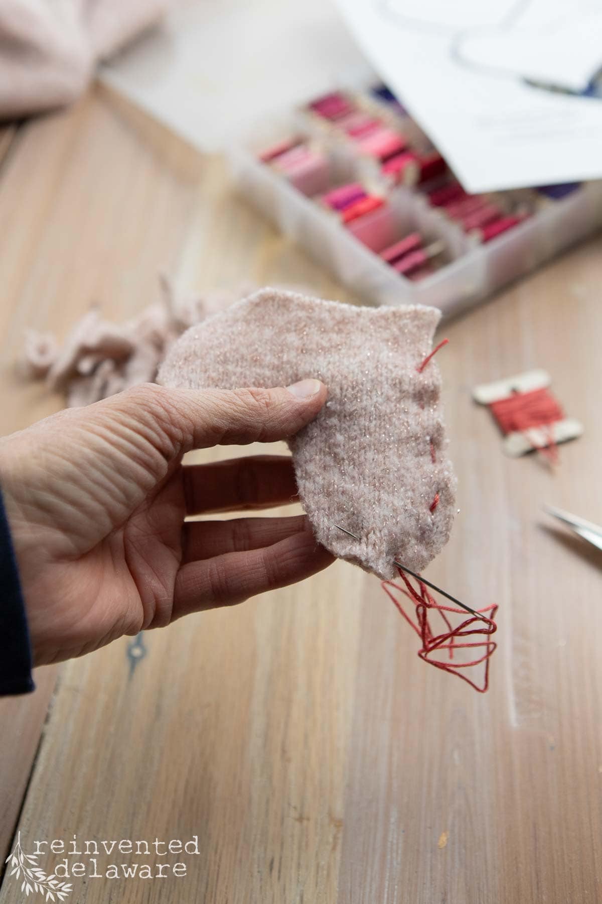 A hand gracefully stitches red thread into a piece of light pink fabric, perfect for DIY Valentine's Day decor ideas. In the background, a box of vibrant spools rests on the wooden surface, inspiring creativity and heartfelt crafts.