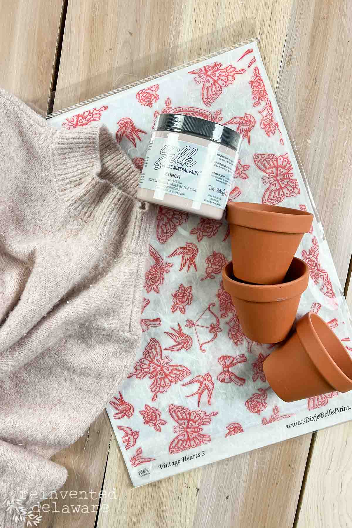 A flat lay showcasing a pink thrift store sweater, a jar of pink chalk mineral paint, and three terracotta pots stacked together, all accented with a red and white decoupage paper featuring floral designs. Perfect inspiration for a Valentine's Day DIY decor project on a wooden surface.