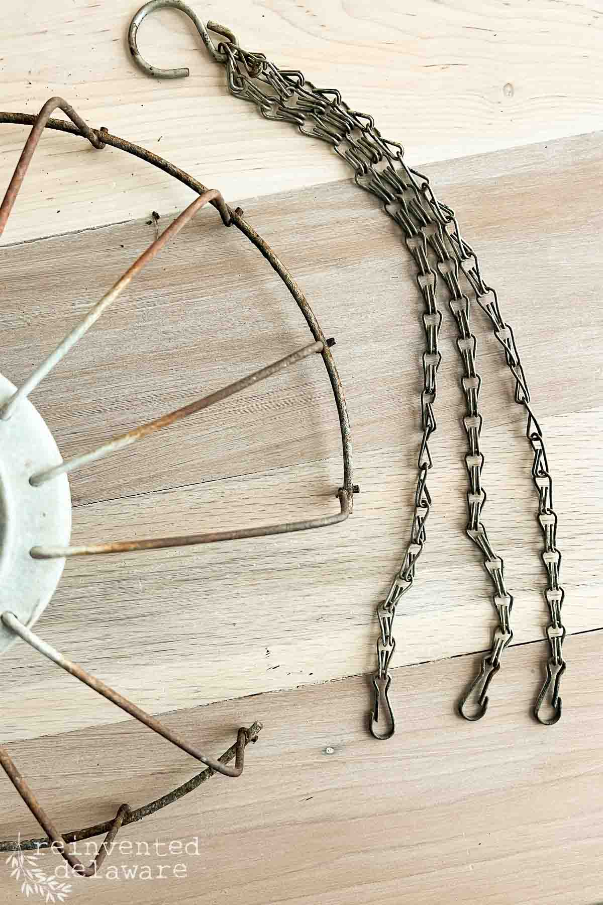 A vintage metal chicken feeder on a wooden surface, featuring a circular frame with aged patina and three metal chains with hooks.
