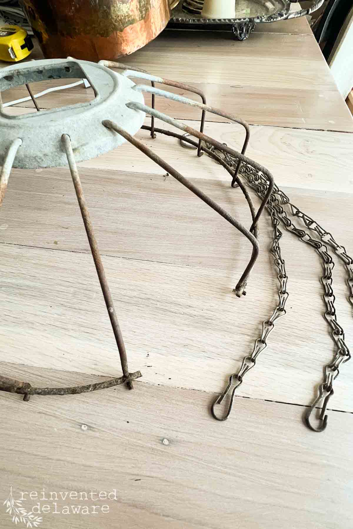 An old, rusted metal chicken feeder with hanging chains sits on a wooden floor. The frame is missing its fabric and is surrounded by rustic decor elements. A "Reinvented Delaware" watermark is visible in the bottom left corner.