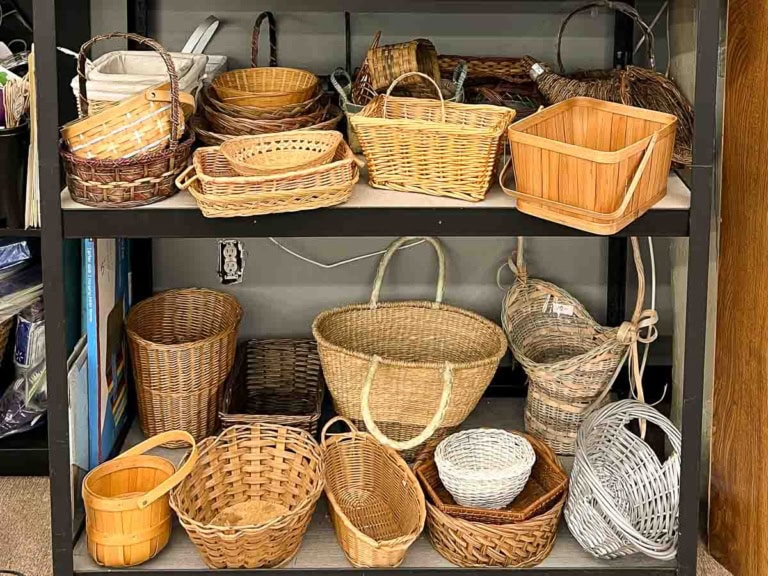 A variety of wicker and wooden baskets are displayed on two shelves, showcasing a thrifting aesthetic perfect for any home decor. The baskets vary in size and shape, with round, square, and rectangular designs. The shelves belong to a storage unit with more items partially visible on the left.