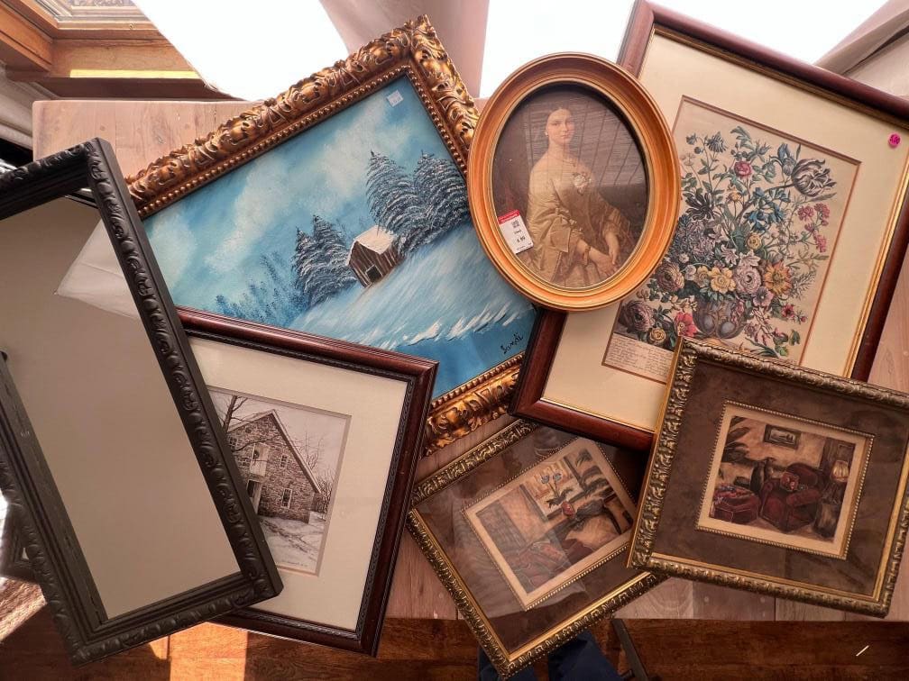 A collection of various framed artworks is displayed on a wooden surface. The frames include paintings and prints, such as a snowy landscape, a floral arrangement, a seated woman, and rustic scenes. The frames vary in size and style.