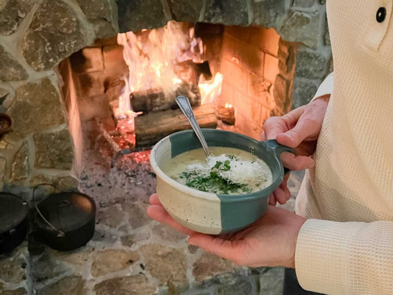 A person holds a ceramic bowl of creamy potato chowder garnished with herbs and grated cheese, standing by a stone fireplace with a fire burning inside.