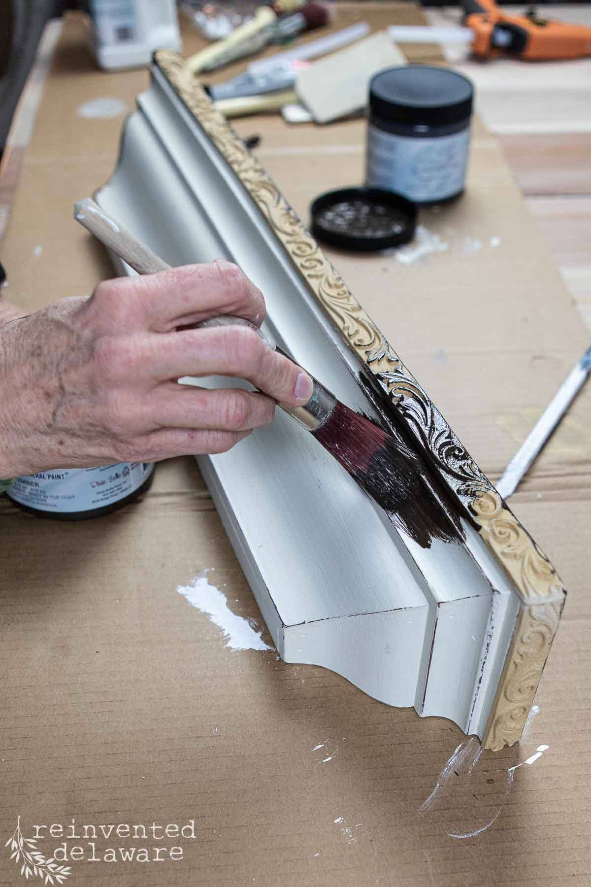 A person is painting an ornate wooden shelf with a small brush. The shelf has intricate carvings on the border. There's a jar of black paint and various tools on the table. The background shows a workshop setting.