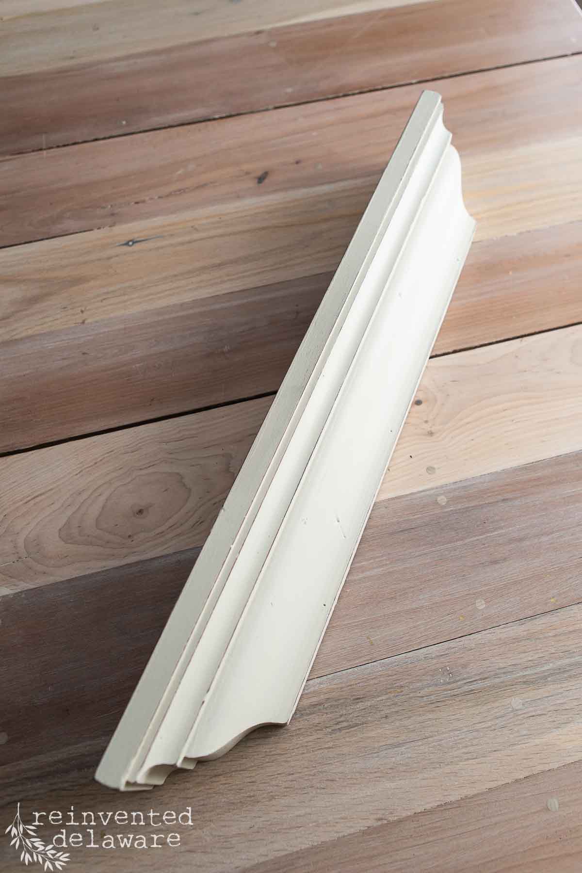 A decorative wall shelf with a creamy white finish graces a natural wood surface, offering the perfect thrift flip wall shelf makeover. The shelf boasts a simple, elegant curved design, while the "reinvented delaware" watermark is subtly visible in the bottom left corner.