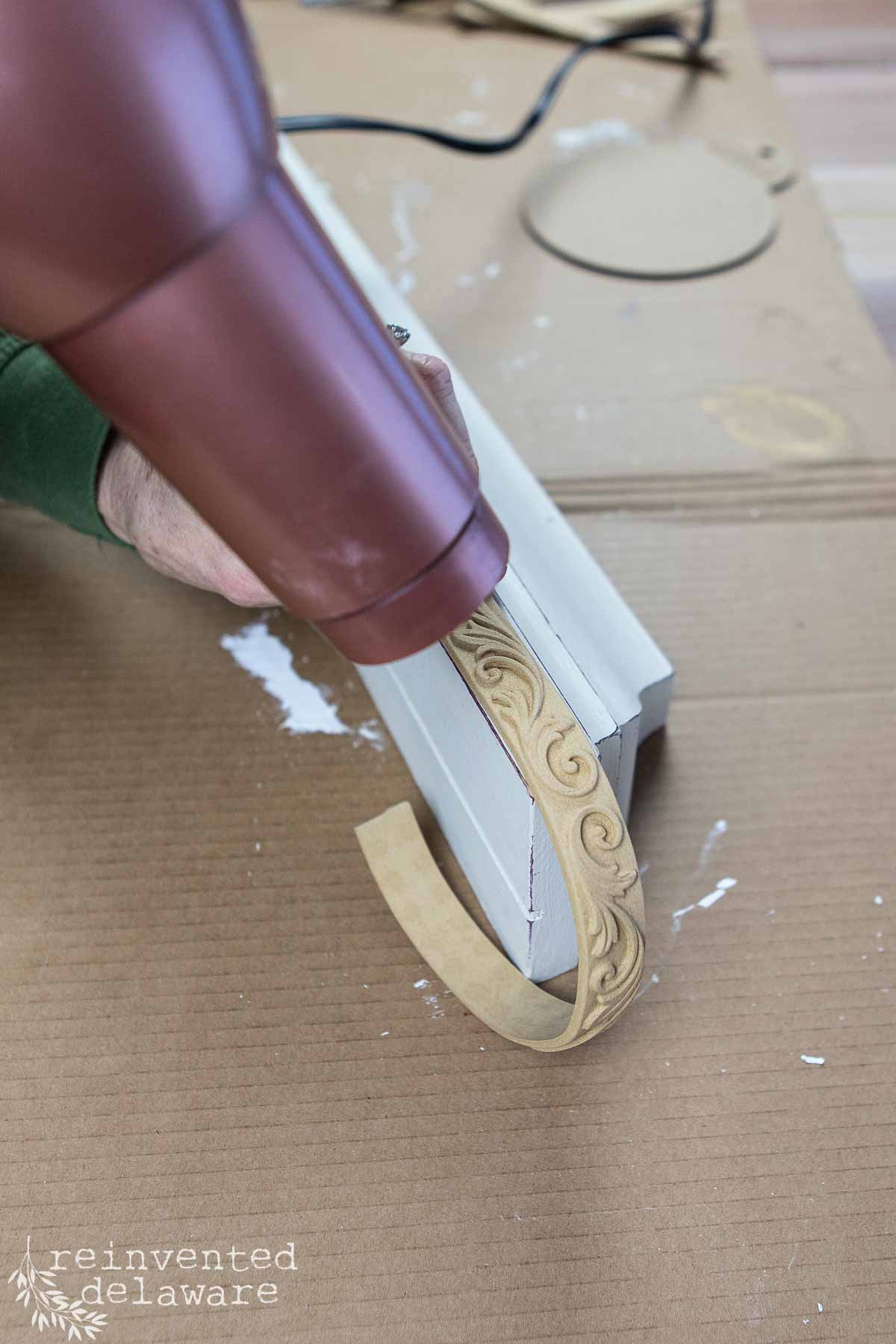 A person uses a hairdryer to apply a decorative wood applique with an intricate swirl design to a white wooden surface on a cardboard mat, transforming it in this creative thrift flip. Tools and materials for the wall shelf makeover are visible in the background.