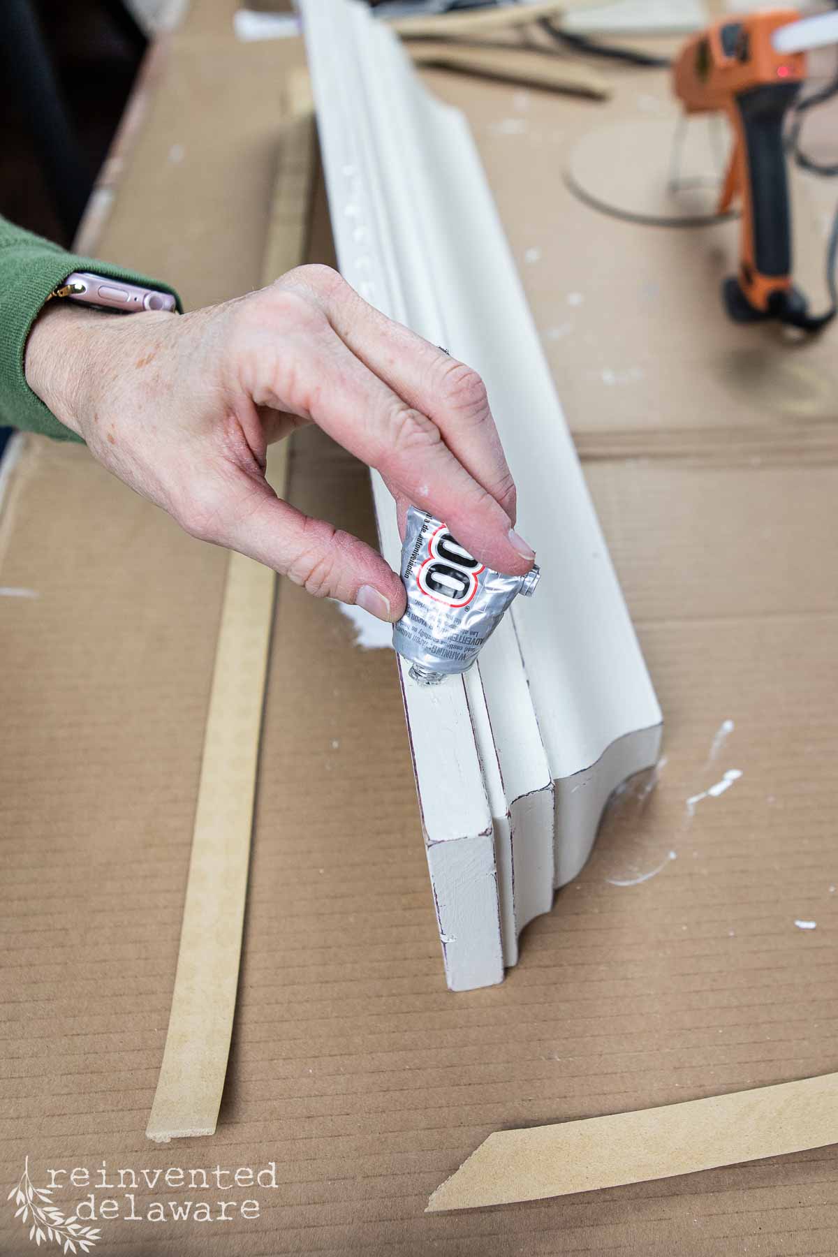 A person in a green sleeve applies glue to intricate decorative molding for a thrift flip wall shelf makeover. The molding, with its decorative grooves, rests on a cardboard surface, surrounded by a glue gun and other tools on the table.