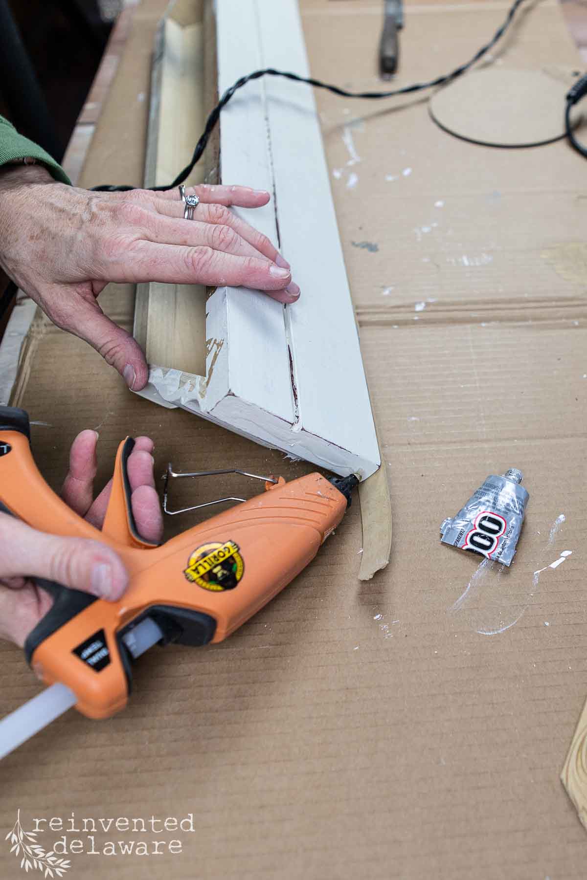 Person using an orange hot glue gun to update a wooden wall shelf on a cardboard surface. A tube of glue and a black cord are nearby. The person wears a green jacket, and the image has "reinvented delaware" in the corner.