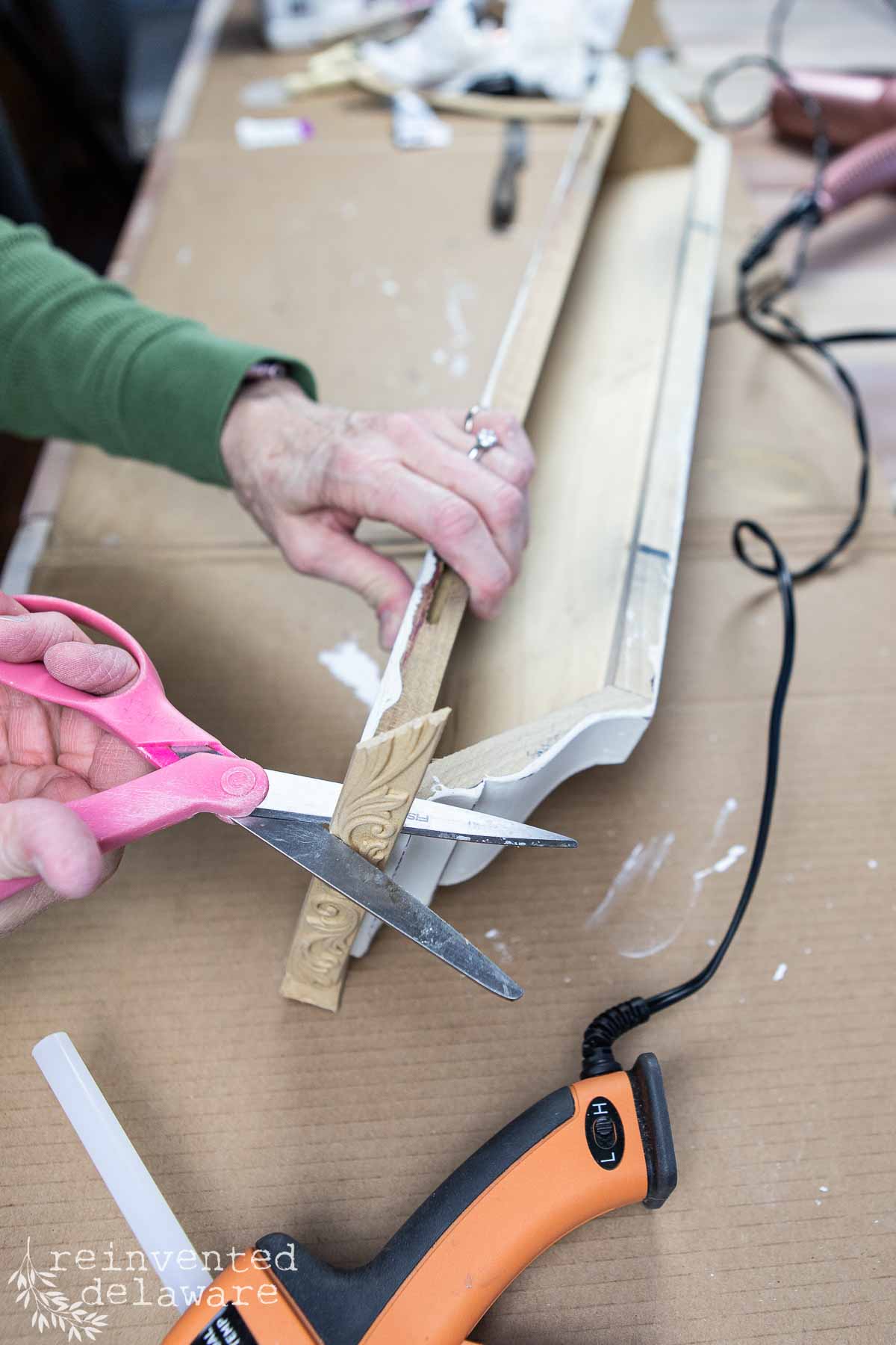 A person in a green shirt uses pink scissors to cut a piece of decorative trim. A wooden wall shelf and a hot glue gun lie on a table covered with cardboard beneath their hands.