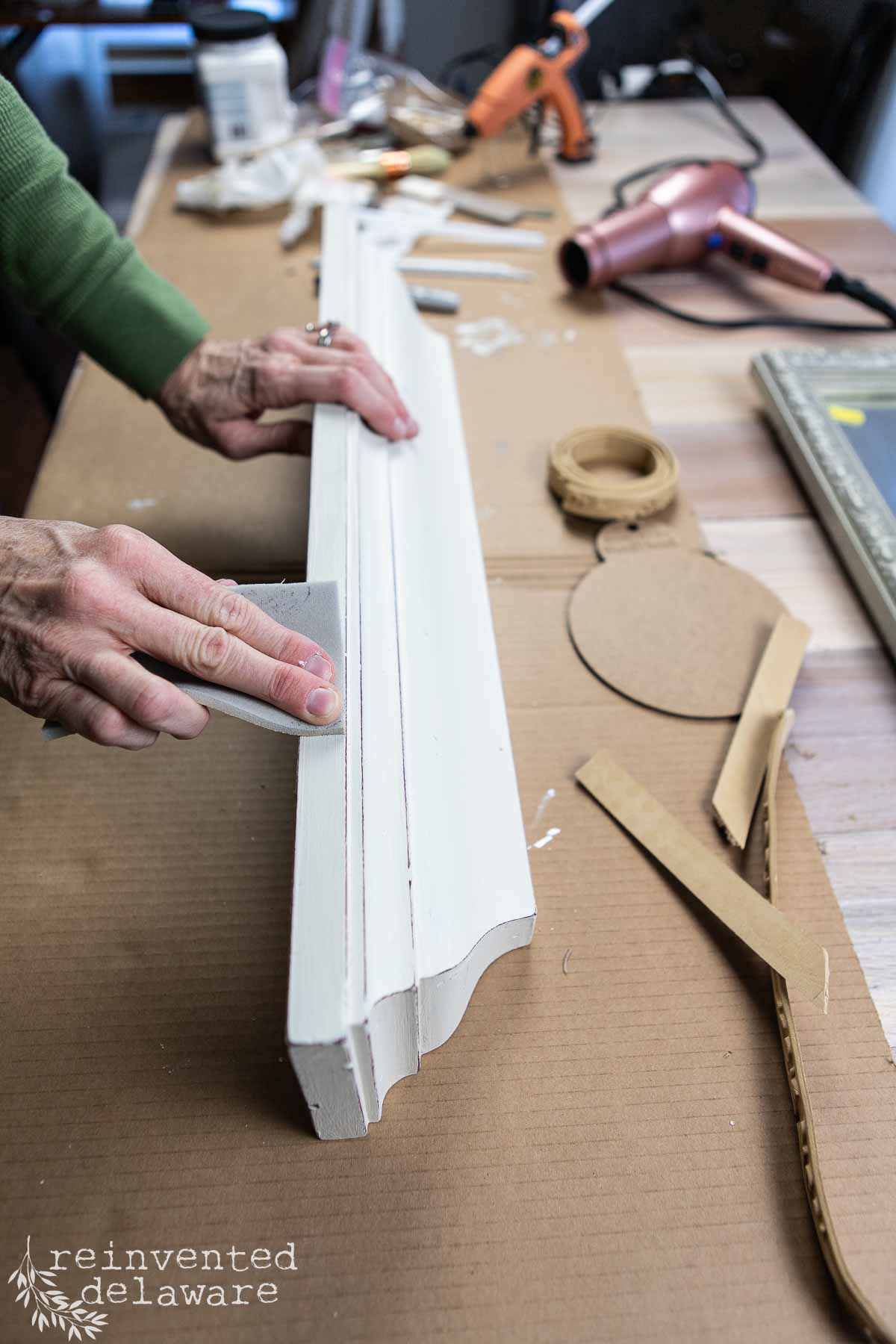 A person sanding a white wooden piece on a cluttered workbench, preparing for a thrift flip wall shelf makeover. Tools like a glue gun and hairdryer lie amidst cut-out cardboard shapes, while the tabletop bears marks of creativity and transformation.