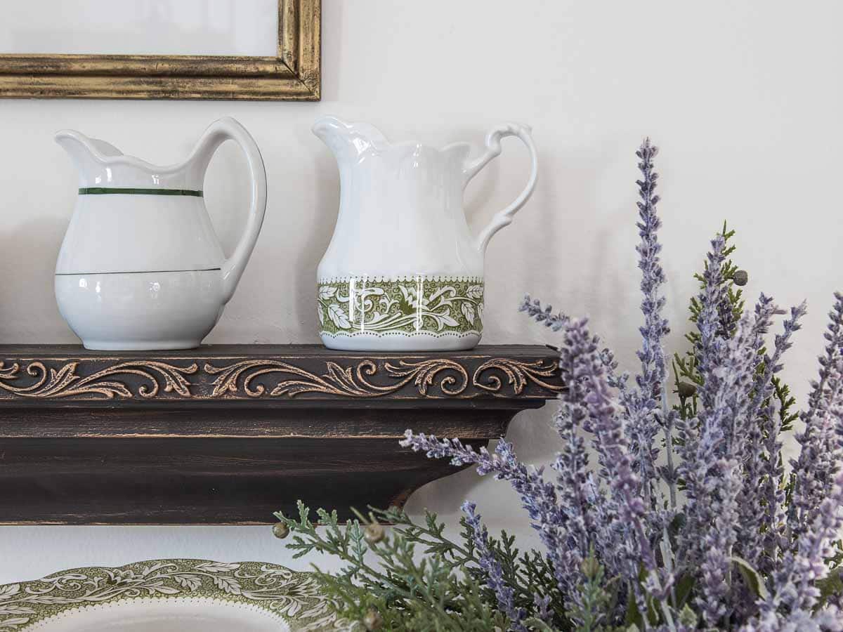 Two ironstone pitchers with green accents sit on an ornate wooden shelf. In the foreground, sprigs of lavender and greenery add a touch of nature. A partial picture frame is visible above.