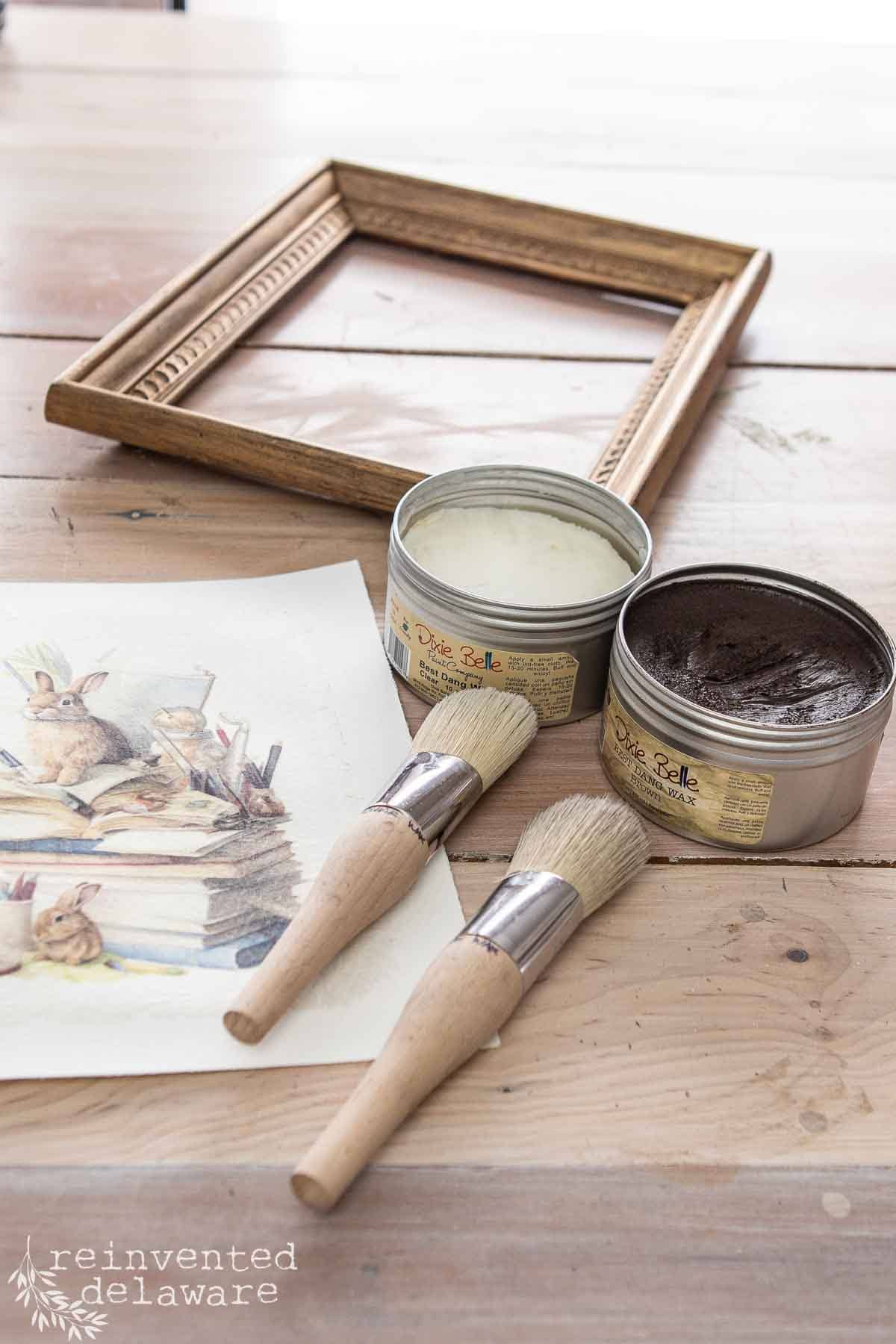 Art supplies on a wooden table, perfect for Easter decorations, include an empty wooden frame, two open cans of cream and dark brown paint, two paintbrushes, and a watercolor painting of rabbits. Ideal DIY ideas for a cozy holiday aesthetic!.