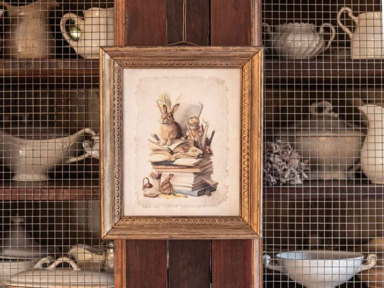 A framed illustration of a rabbit, perfect for Easter decorations, sits atop a stack of books on the wooden cabinet door. Behind the screen door, white ceramic dinnerware and teapots are arranged on shelves.