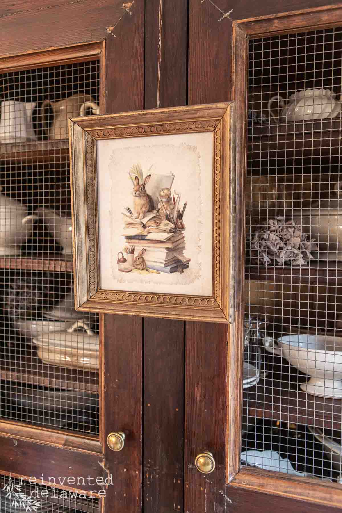 A framed picture of rabbits resting on books adorns the doors of the dark wooden cabinet with mesh panels, adding a touch of Easter charm. Inside, white ceramic dishes and a decorative item are visible, offering inspiration for your own DIY ideas.