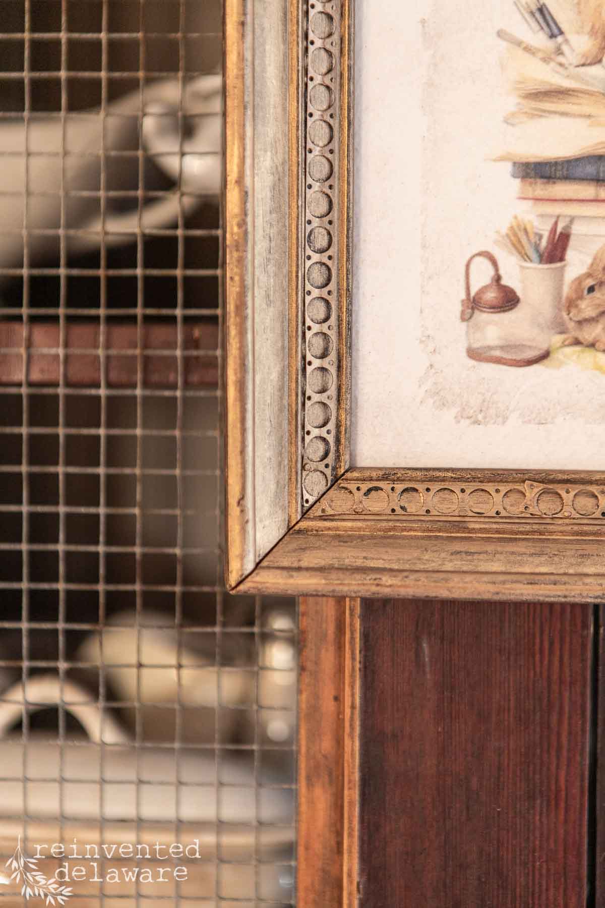 Close-up of a vintage wooden frame with a decorative border, displaying part of an artwork featuring books and an oil lamp. The frame is against a wire-mesh cabinet containing dishes and subtle Easter decorations. Branding text "reinvented delaware" is visible in the corner.